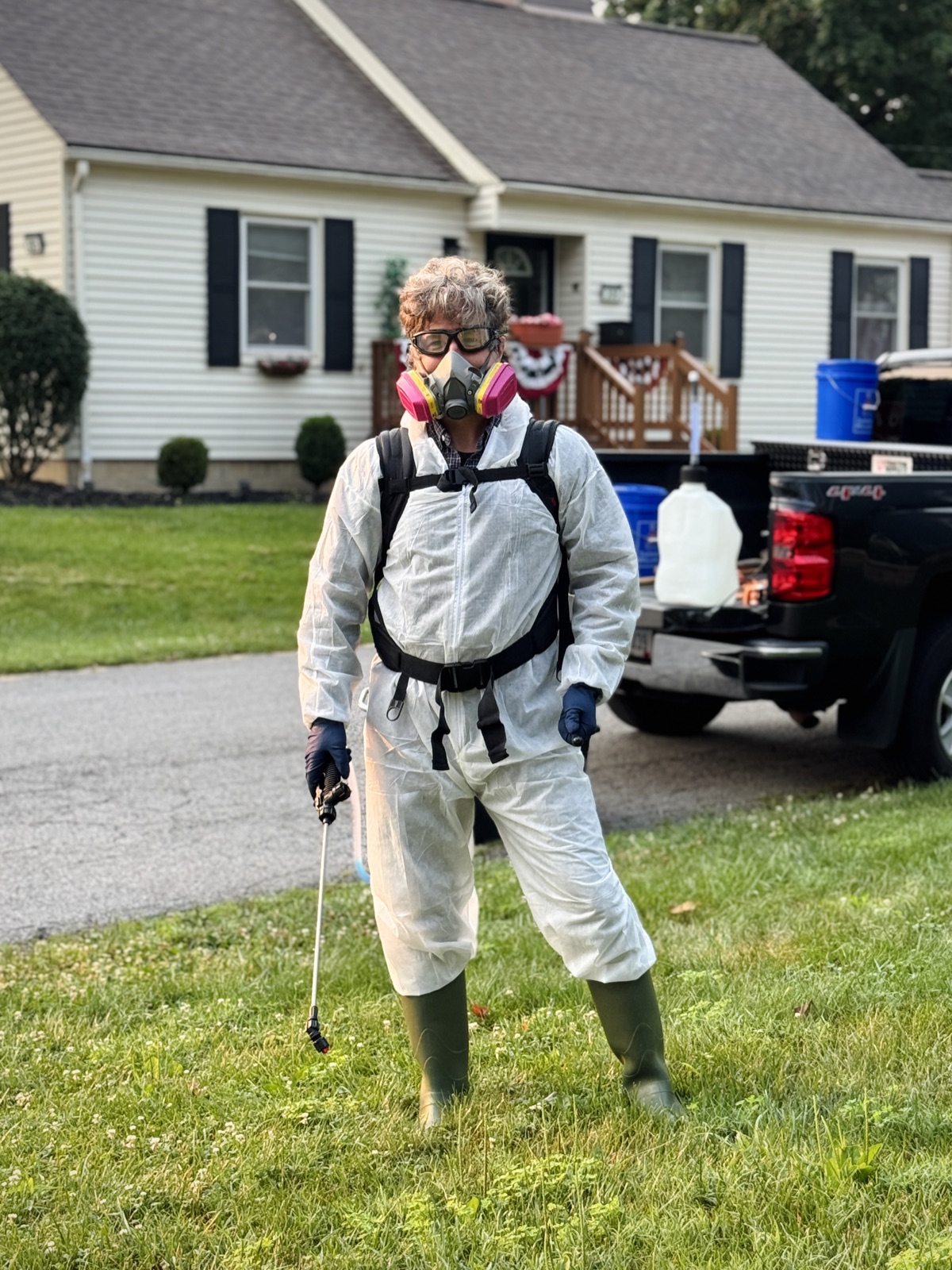 David All in full protective coverall, respirator, goggles and gloves, holding a spray wand, with a treatment tank in the truck bed behind him.